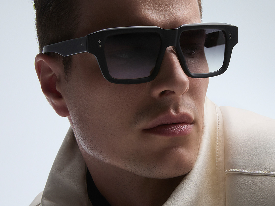 Man wearing large black sunglasses and a light-colored jacket against a plain background. “DITA” is written across the bottom in white text.
