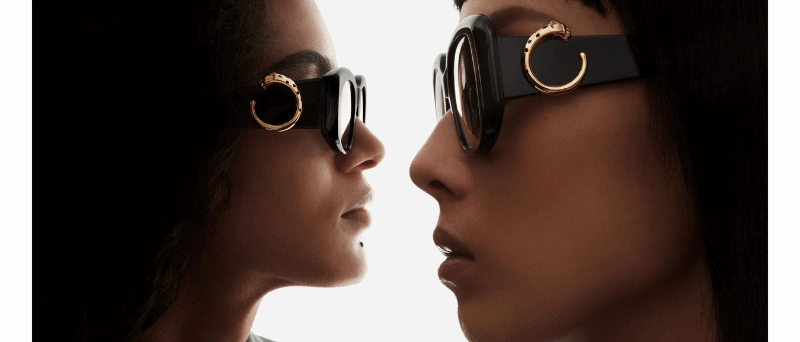 Two women in profile face each other, both wearing dark, round sunglasses with large gold decorative accents.