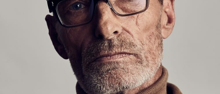 An older man with light brown hair, wearing black glasses, a brown turtleneck, and a matching brown blazer, looks directly at the camera against a neutral background.