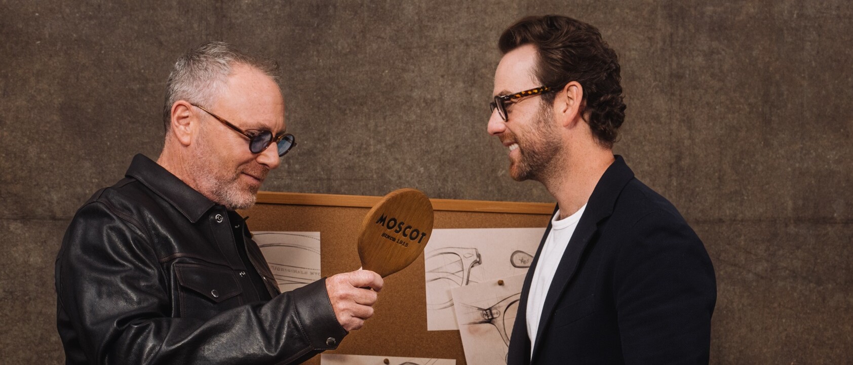 Two men wearing glasses stand in front of a corkboard with sketches; one holds a wooden paddle labeled 