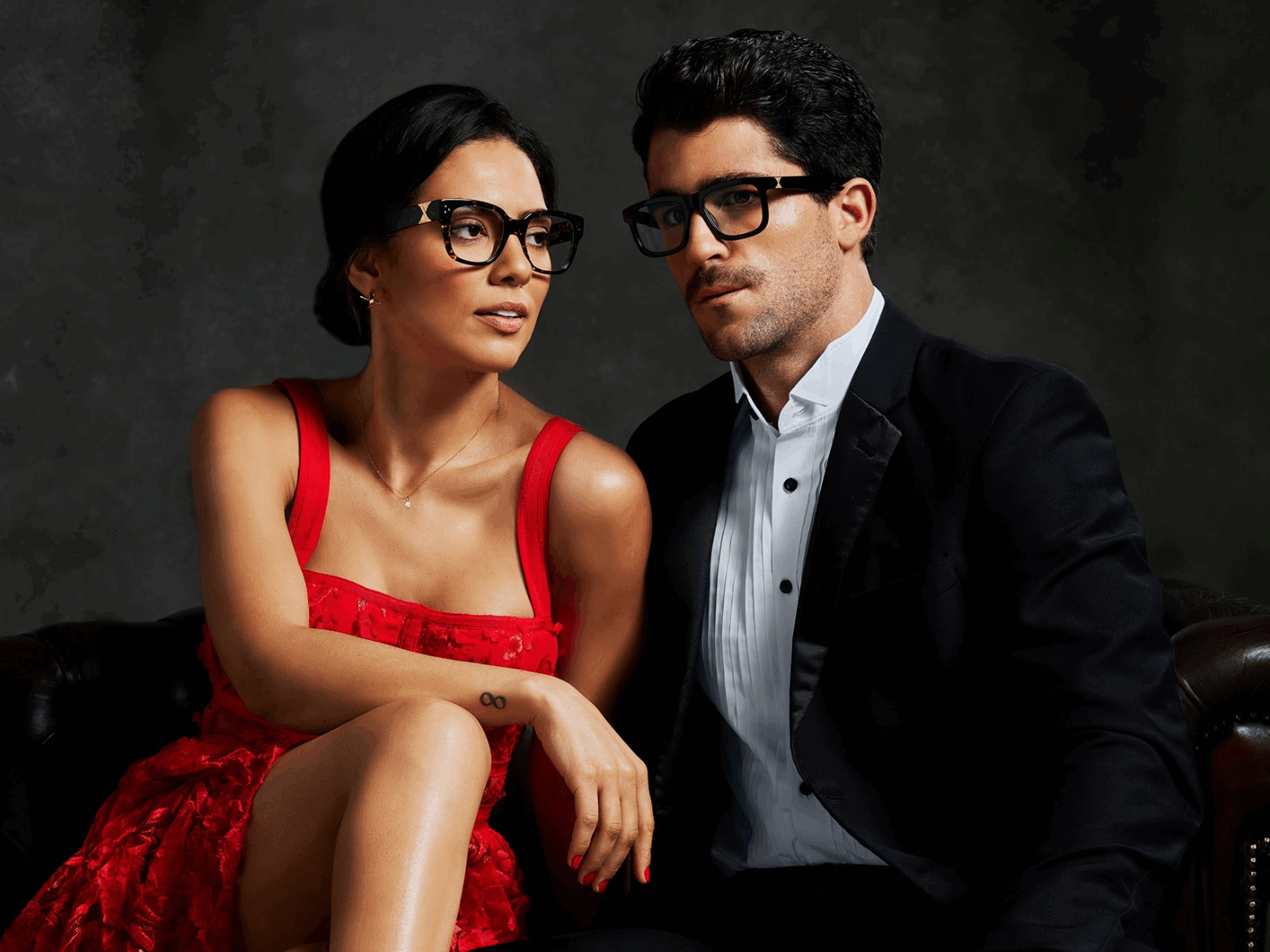 A woman in a red dress and a man in a black suit and glasses sit together on a dark leather sofa against a dark background.