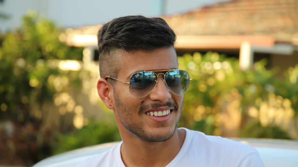 A man wearing sunglasses and a white t-shirt smiles outdoors, with trees and a building in the blurred background.