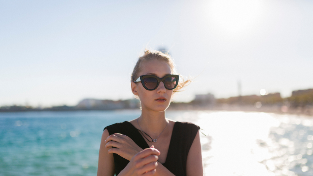 A woman wearing sunglasses stands by the water on a sunny day, with buildings and a bright sky visible in the background.
