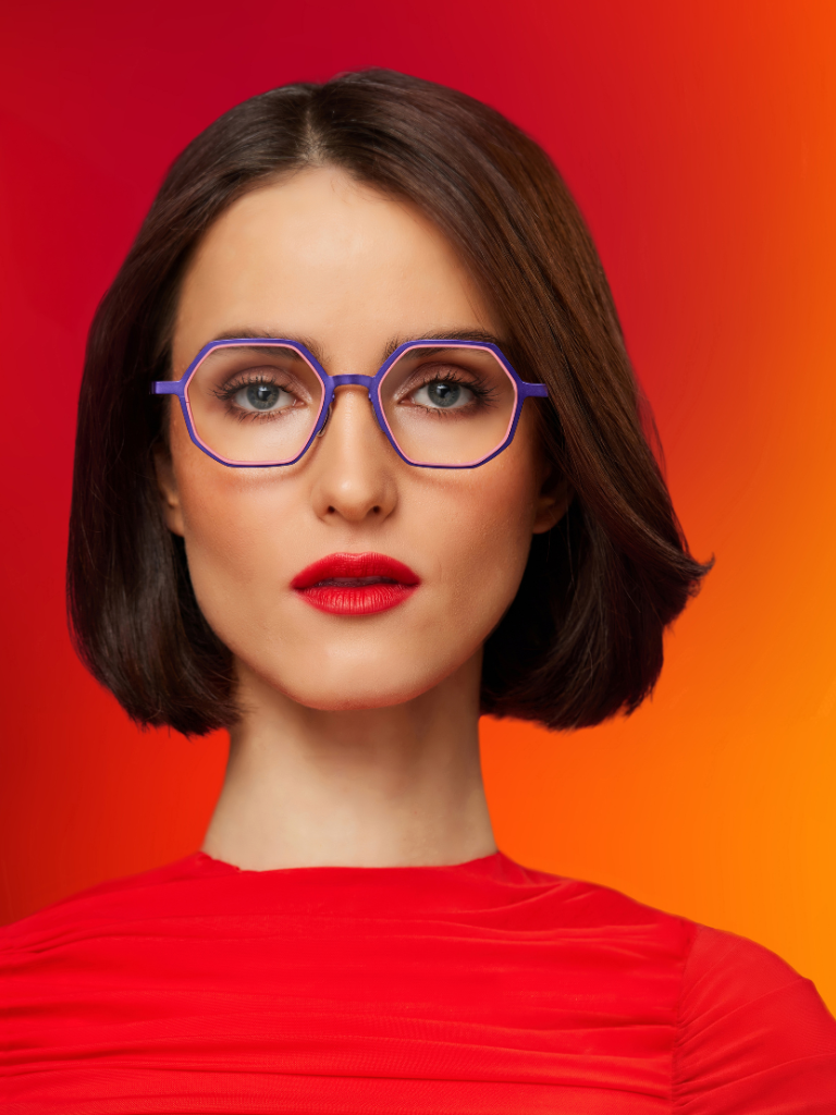 Woman with short brown hair, wearing purple glasses and a red top, poses against a red and orange gradient background.
