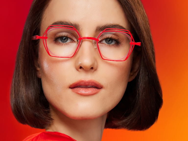Woman with straight brown hair wears red-framed glasses and an orange top. Text below reads: