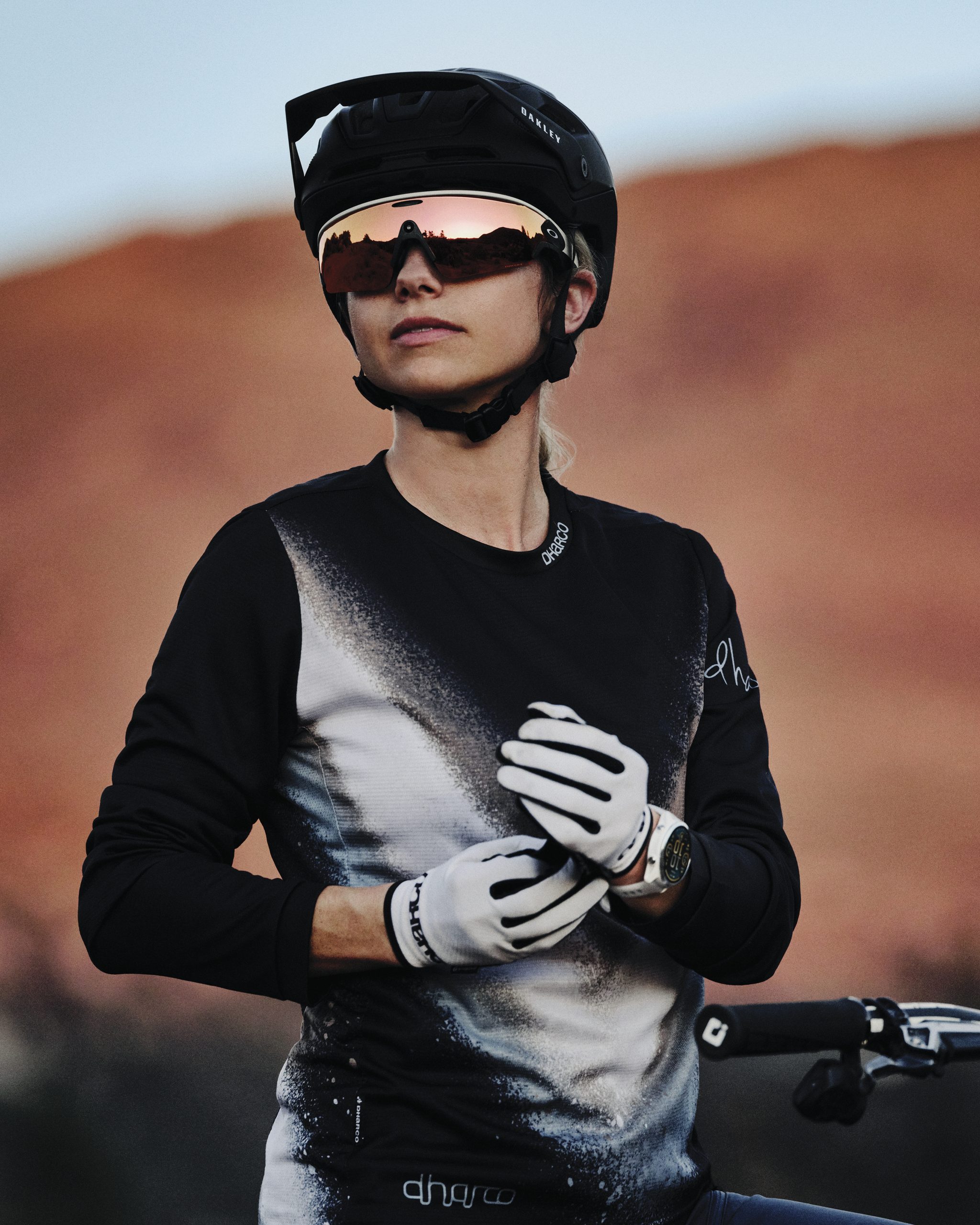 Person wearing a black helmet, Ray-Ban Meta reflective sunglasses, gloves, and a long-sleeve shirt stands next to a bicycle with a red rocky landscape in the background.