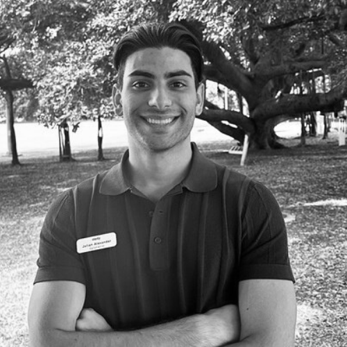 A man with his arms crossed wearing a name tag that reads “Julian Alexander” in a park.