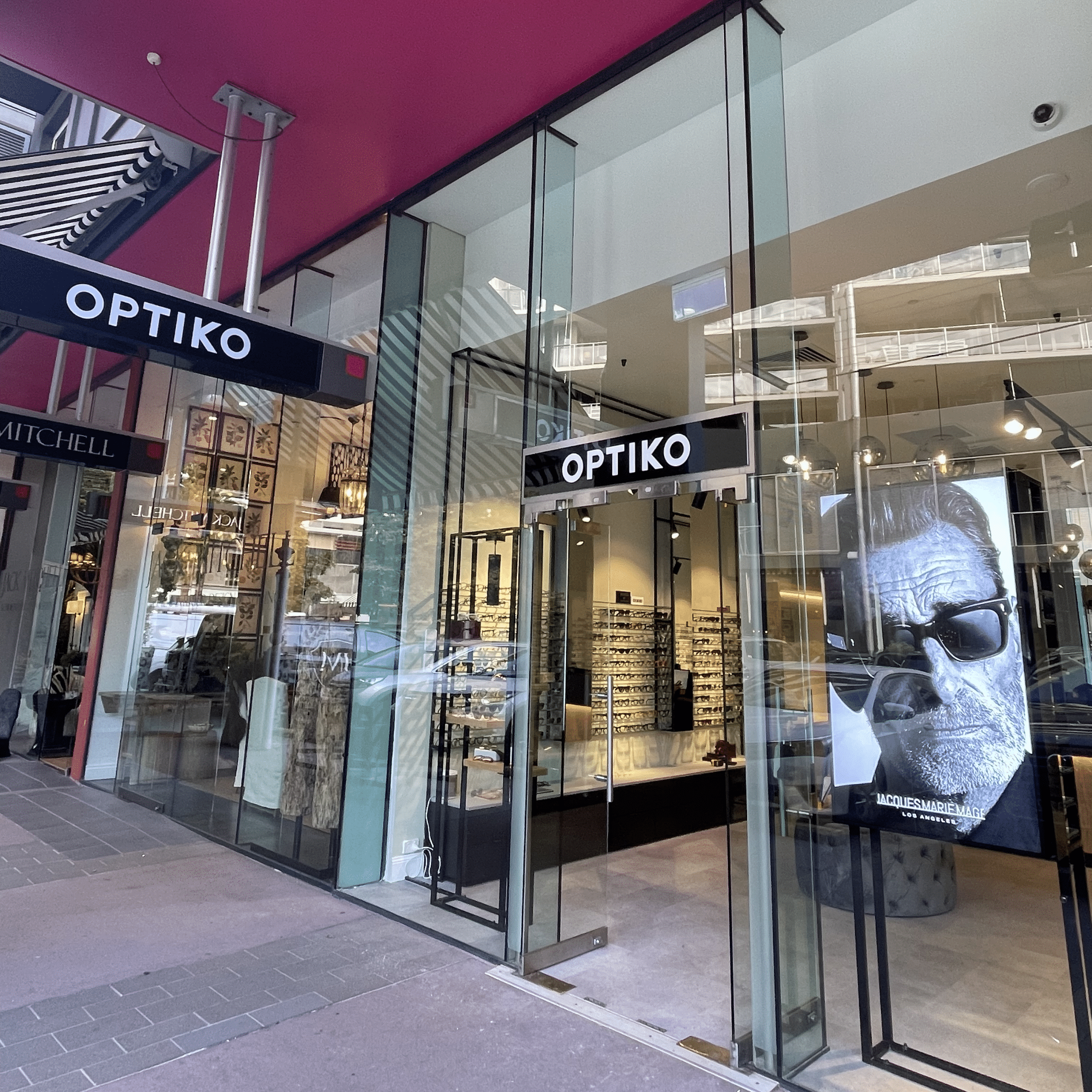 The storefront of an Optiko eyewear shop with glass doors and a large poster of a man wearing sunglasses in the window.