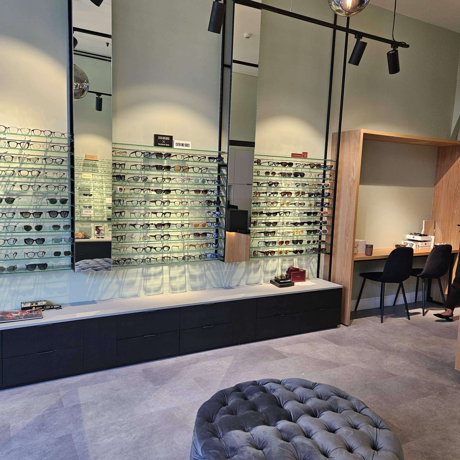 Interior of an optical shop with wall-mounted shelves displaying rows of eyeglasses, a central tufted grey ottoman, and a desk with two chairs.