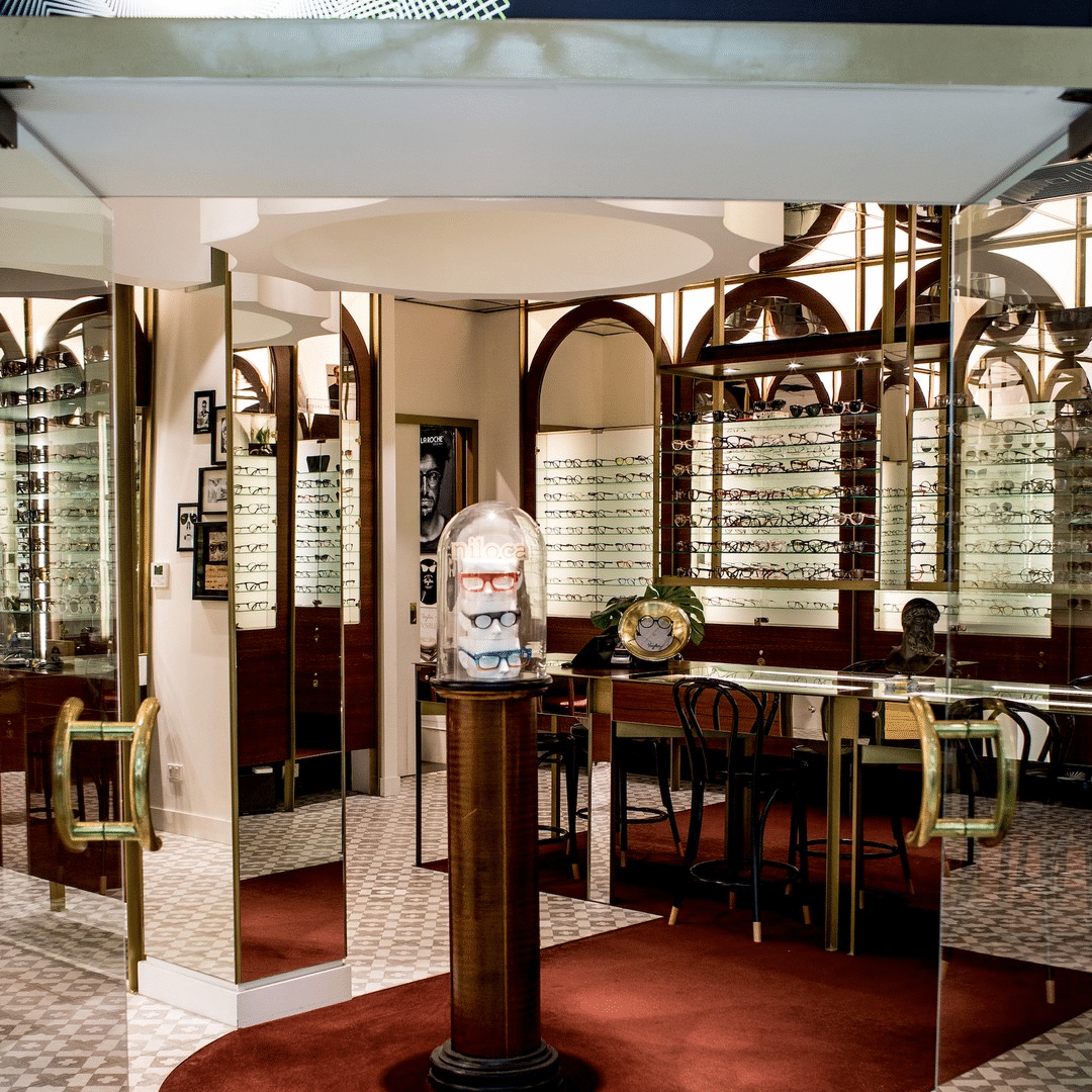 Interior view of an Optiko eyewear shop featuring rows of eyeglasses on display and a pedestal with a glass dome showcasing a stack of three pairs of glasses.