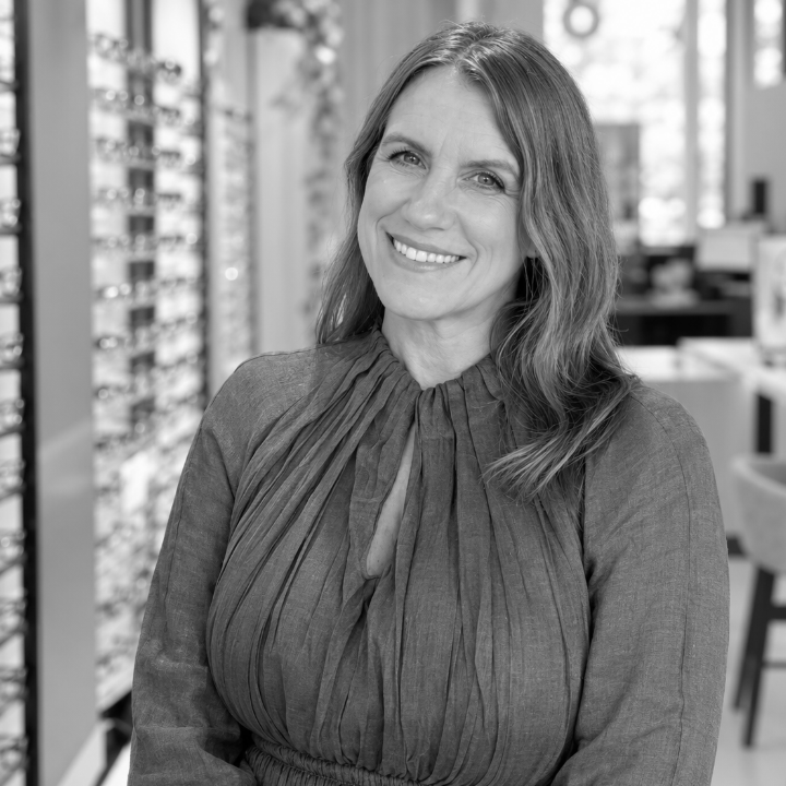 A smiling woman standing in an optical shop with displays of glasses in the background.